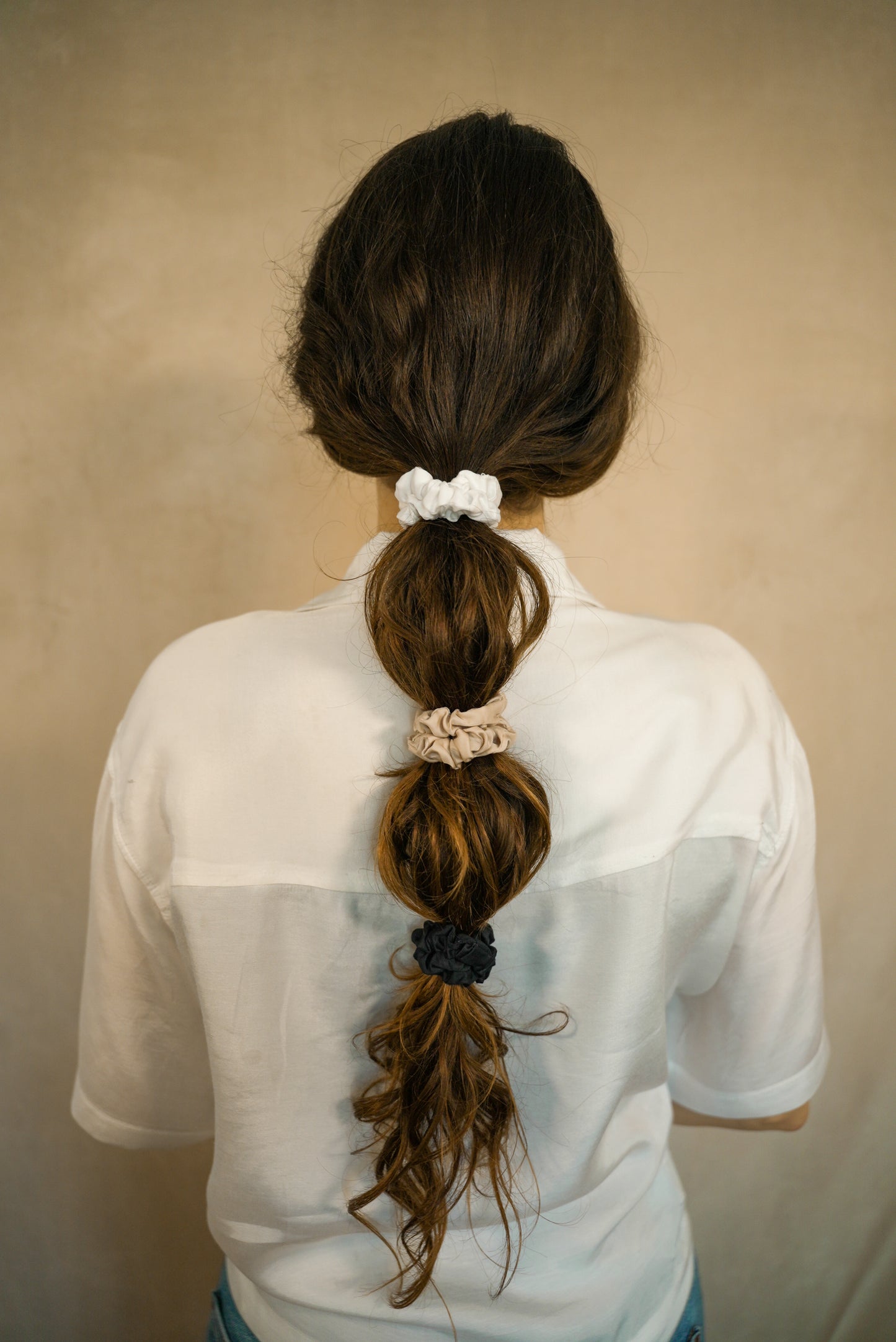 Curly girl with a ponytail and 3 scrunchies wearing a white shirt against a beige background